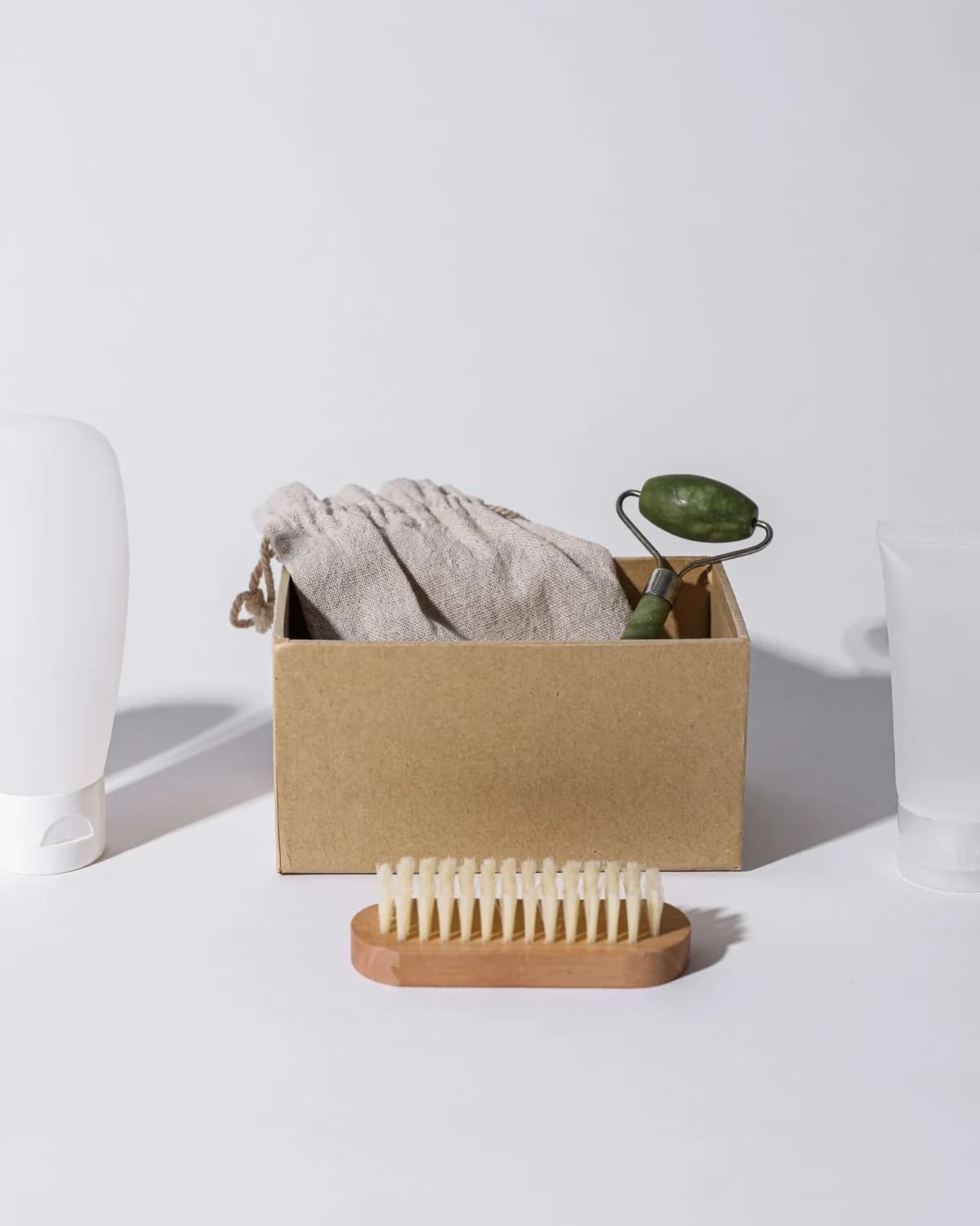 Skincare tubes, a jade roller and a wooden brush arranged with a kraft box on a pale ground